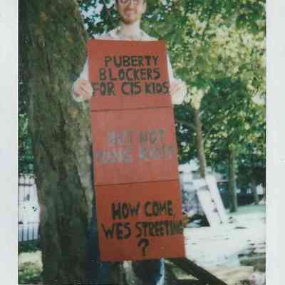A person stands by a tree holding a sign which says "Puberty blockers for cis kids but not trans kids. How come Wes Streeting?"
