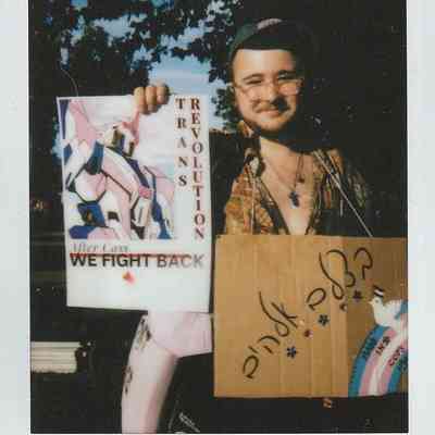 A person holds two signs expressing themes of trans pride and activism while smiling outdoors.
