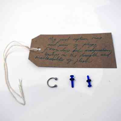 A handwritten tag, metal septum ring, and blue plugs are displayed on a white surface.