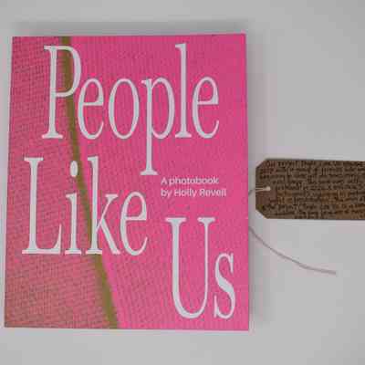 The image features a photobook titled "People Like Us" by Holly Revell, which has a pink cover with a green line through it. Next to it is a brown tag with writing on it.