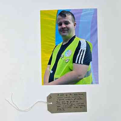 A person wearing a yellow vest with "Trans Pride" stands against a colorful background and a handwritten note