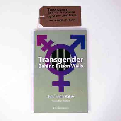 A book called 'Transgender Behind Prison Walls' by Sarah Jane Baker which has a grey background and the transgender symbol in a gradient pink and purple hue.