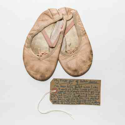 Pink ballet shoes tied together with a small piece of white string. Both shoes have black marks on the bottom and show other signs of wear such as dirty elastic over each bridge and frayed seams.