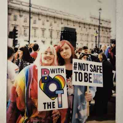 Two people holding colorful protest signs which say "B WITH THE T" and "#NOT SAFE TO BE ME".