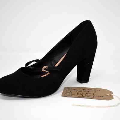The image shows a black high-heeled shoe with a strap, accompanied by a handwritten tag.