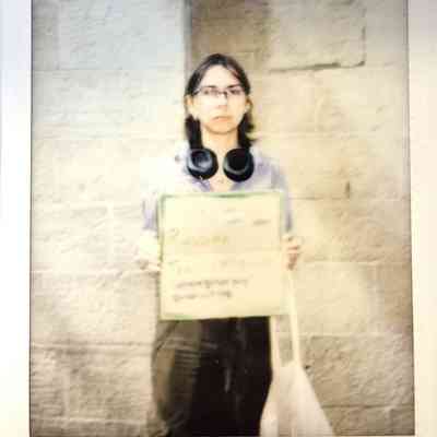 A person standing against a brick wall holding a handwritten sign, with headphones around their neck.