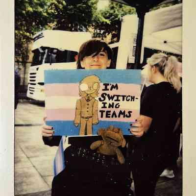 A person is smiling while holding a sign with a drawing and the text "I'M SWITCHING TEAMS".