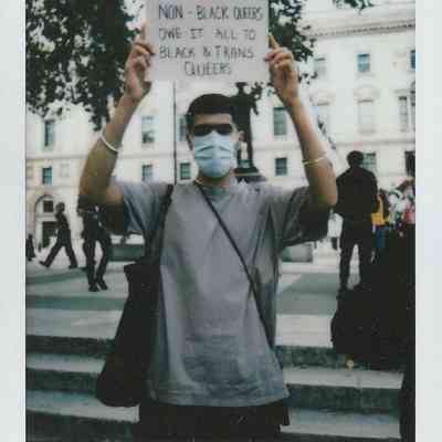 A person wearing a mask holds a sign which says "Non-Black Queers Owe It All To Black & Trans Queers".