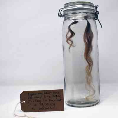 A clear glass Mason Jar containing two locks of brown, human hair, suspended on thread. One lock is longer than the other. With a brown, handwritten luggage tag.