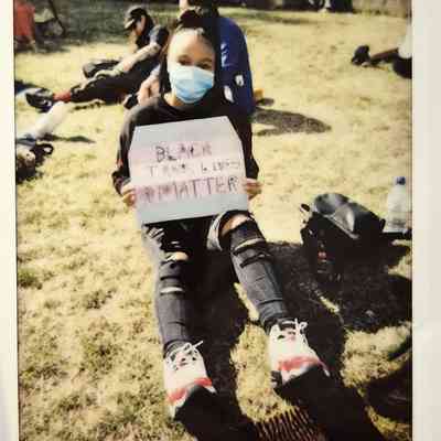 A person sits outdoors, wearing a mask, holding a sign supporting Black trans lives.