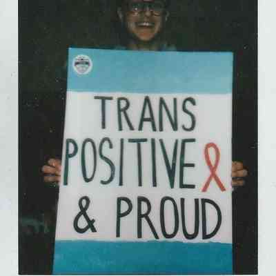 A person is smiling while holding a sign that says, "Trans Positive & Proud," in bold text.