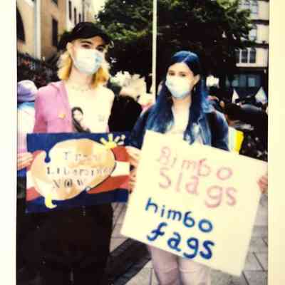 Two people with masks hold colorful signs at a public gathering.