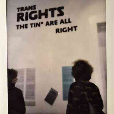 The image shows two people looking at a wall with a sign reading "Trans Rights The Tin* Are All Right".