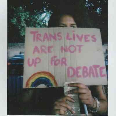 A person holds a cardboard sign stating, "Trans lives are not up for debate," featuring a rainbow.