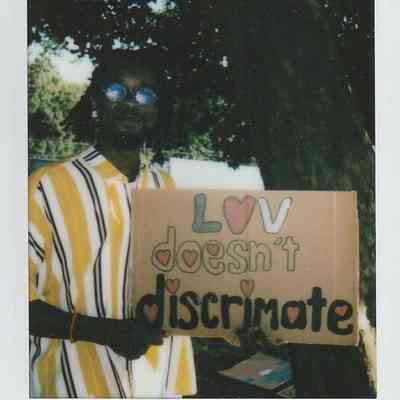 A person stands holding a cardboard sign that says, "Love doesn't discriminate," under a tree.