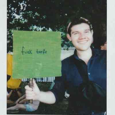A smiling person is holding a sign with text in a park, standing against a backdrop of trees.