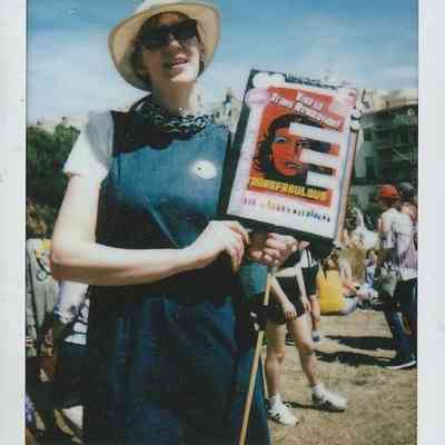 A person in sunglasses and a hat holds a sign which says "Viva La Trans Revolution!".