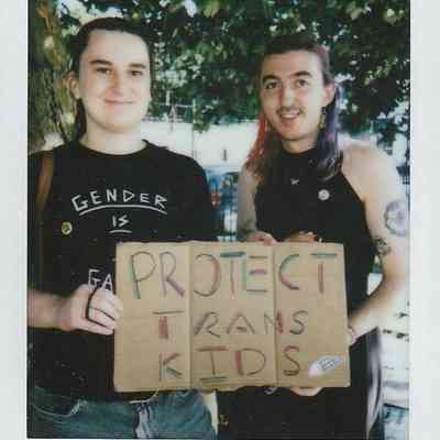 Two people are smiling while holding a cardboard sign that says "Protect Trans Kids" in colorful letters.