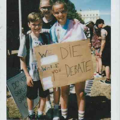 Three people stand together at an outdoor event, holding a sign which says "We Die While You Debate".