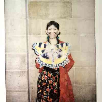 A person stands against a wall, wearing colorful face paint and holding a butterfly-shaped placard.