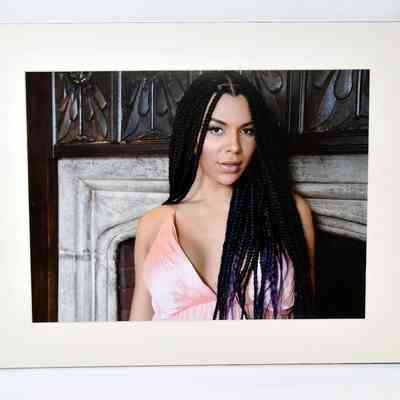 Colour photo of woman with long dark hair braids in pink dress standing in front of fireplace.