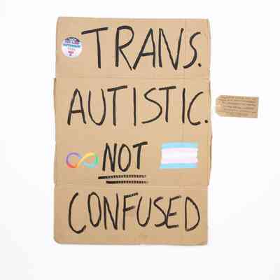 Handwritten cardboard placard reads: "TRANS. AUTISTIC. NOT CONFUSED," ["NOT" is double underlined] with colorful symbols. Decorated with a sticker, a rainbow colored infinity symbol and the trans flag. With a brown, handwritten luggage tag.