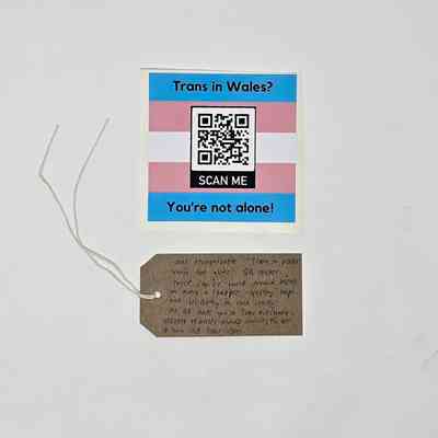 Square sticker in the trans flag colours. There is a QR code in the center and the text "Trans in Wales?" above this and  "You're not alone!" below. Also, a brown hand written luggage tag
