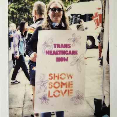 A person smiles while holding a sign advocating for transgender healthcare, surrounded by people on the street.