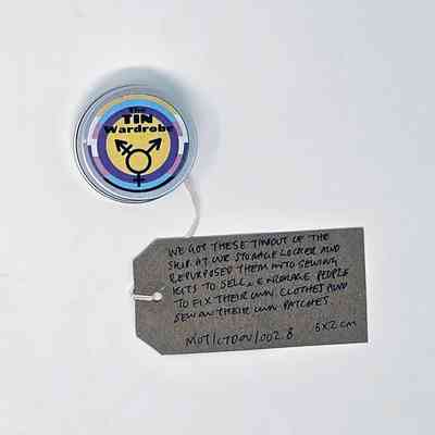 A small, round, silver tin labeled "The TIN Wardrobe". The label is in the trans, non-binary and intersex pride flag colours and features the trans symbol. With a brown handwritten luggage tag.