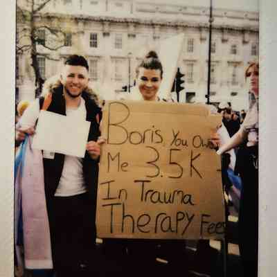 Two people in a protest hold a cardboard sign which says "Boris, you owe me 3.5k in trauma therapy fees".