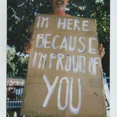 A smiling person holds a supportive sign reading, "I’m here because I’m proud of you,"