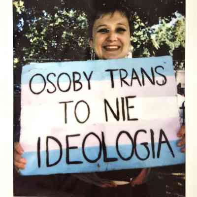A person holds a sign reading "Osoby Trans to nie ideologia" with a smile outdoors.