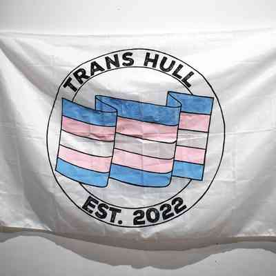 A large white banner with two concentric circles containing the text "TRANS HULL" and "EST. 2022" at the top and bottom. Side the circles is a rippling blue, white and pink trans pride flag
