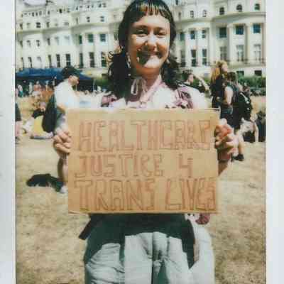 A person smiles holding a sign that reads "Healthcare Justice 4 Trans Lives" at an outdoor gathering.