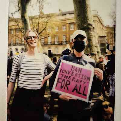 Two people are standing outdoors, holding a sign that says, "Ban Conversion Therapy for All."