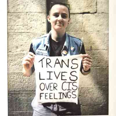 A person stands against a stone wall, holding a sign that reads, "TRANS LIVES OVER CIS FEELINGS".
