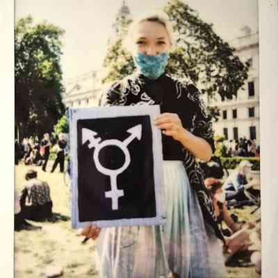 A person wearing a mask holds a sign with a gender symbol at an outdoor event.