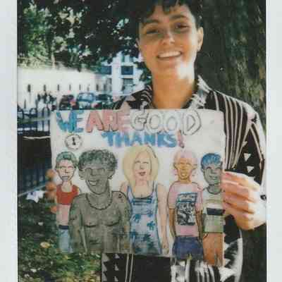 A person smiles while holding a colorful drawing reading, "We are good thanks!" in an outdoor setting