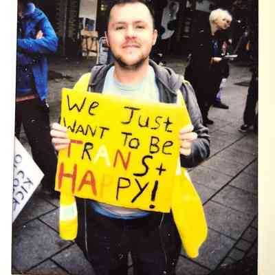 A person holds a bright yellow sign reading "We just want to be trans + happy!"