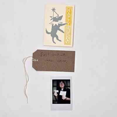 A yellow birthday card depicting a grey dog wearing a ruff and a pointy hat. With a brown, handwritten luggage tag and a polaroid of a person holding the card.