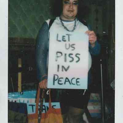 A person is holding a sign advocating for bathroom rights, stating, "LET US PISS IN PEACE".