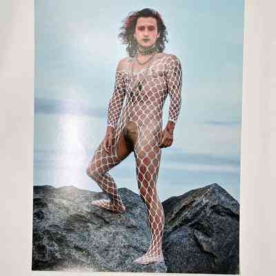 A person who is wearing a white fishnet bodysuit and is stood on rocks in front of the ocean.