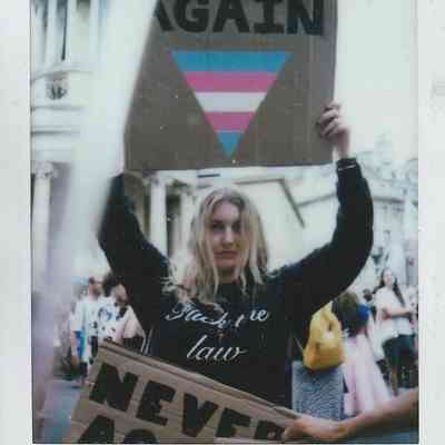 A person holds a cardboard sign which says "Never Again" with an upside-down triangle painted in trans flag colours.