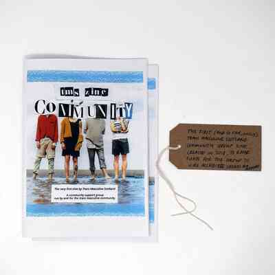 Two copies of the Trans Masculine Scotland (TMS) Community Group zine. Cover image is 4 people paddling in water with the title obscuring their faces. With a brown handwritten luggage tag.
