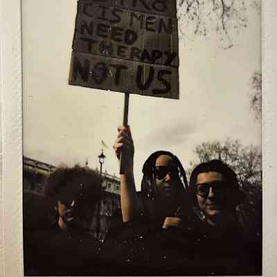 Three individuals stand together, holding a protest sign that reads, "STR8 CIS MEN NEED THERAPY NOT US".
