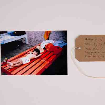 A young, dark haired child wearing white shorts lies on a red, slatted, wooden sunbed on a sandy beach. Their legs are crossed at the ankles and their hands are behind their head. With a brown, handwritten luggage tag.