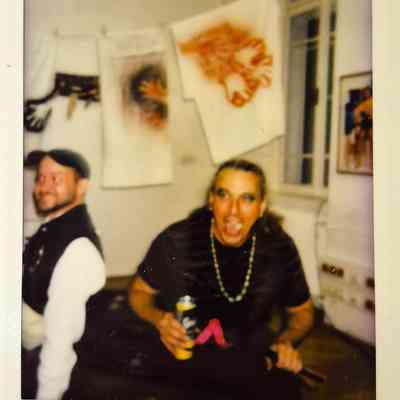 Two people are in a room with colorful art and a beverage.