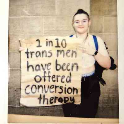 A person is holding a sign reading, "1 in 10 trans men have been offered conversion therapy".
