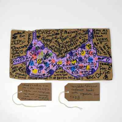 A drawing of a light purple bra, with darker purple spots, on a piece of brown cardboard. It has been signed my many different people. With two brown, handwritten luggage tags.
