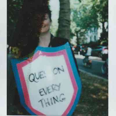 A person partially obscured by a shadow holds a shield-shaped sign saying "QUESTION EVERYTHING" outdoors.